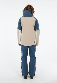 Waterdichte jas in beige en blauw met een aansluitende capuchon, contrasterende panelen en een langere achterkant. Gecombineerd met blauwe skibroeken en ritsen.