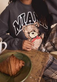 Personne portant un sweat-shirt foncé avec "MIAMI" et un dessin d'ours en peluche, assise à une table en bois avec un croissant sur une assiette verte.