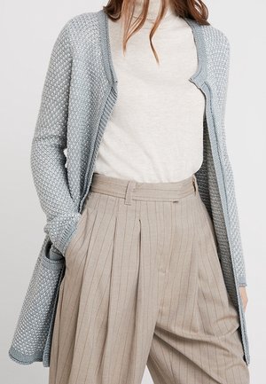 Kvinde iført en lysegrå tekstureret cardigan over en hvid rullekrave, kombineret med højhælede beige plissee bukser med striber.