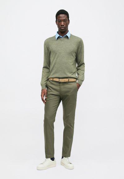 Un giovane uomo sta in piedi indossando un maglione verde chiaro sopra una camicia a colletto blu, pantaloni oliva con una cintura beige e sneakers bianche su uno sfondo bianco.