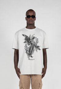 Light gray t-shirt featuring a black and white angel graphic with stylized text below. Short sleeves and relaxed fit. Material appears soft.