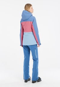 Giacca da sci blu e rosa con cappuccio, caratterizzata da un design a blocchi di colore. Abbinata a pantaloni da sci blu e stivali da sci neri.