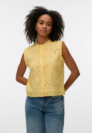 Kobieta z kręconymi włosami, ubrana w żółtą bezrękawnikową bluzkę z ażurowej koronki i niebieskie dżinsy, stoi ze złożonymi za plecami rękami na białym tle.