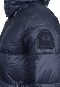 Námořnická modrá péřová bunda s prošívaným designem, která má kapuci a texturovanou nášivku loga na rukávu. Hladká, lesklá látka.