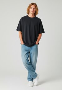 Juodas trumpa rankovė t-shirt, laisvas kirpimas, derinamas su šviesiai mėlynomis džinsų kelnėmis ir baltomis sportinėmis batelėmis, prieš paprastą šviesų foną.