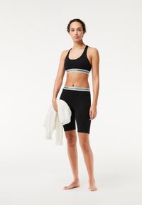 Completo sportivo nero composto da un reggiseno corto e pantaloni aderenti, con bande elastiche a righe bianche e verdi. Il modello tiene una felpa bianca.