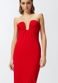Crvena haljina bez naramenica s dubokim V-izrezom i naboranim detaljima na grudima. Glatka tkanina, uski silueta, doseže do sredine lista.