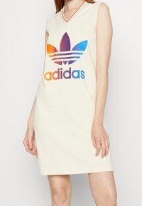 Kremowa sukienka bez rękawów z dekoltem w kształcie litery V, z wielokolorowym logo Adidasa oraz wzorem trzech pasków w kolorach gradientowych. Miękka tekstura materiału.