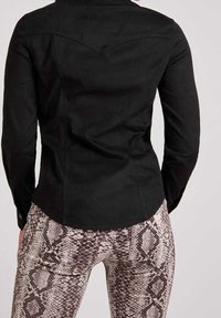 Camicia in suede nero con maniche lunghe, orlo curvo e dettagli sul retro, abbinata a leggings con motivo a serpente in tonalità beige e marrone.