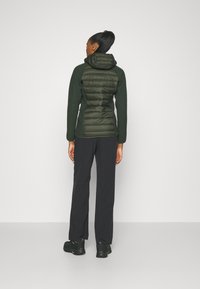 Green puffer jacket with a fitted design, textured fabric, and hood. Paired with black pants and sturdy black footwear. Visible stitching details.