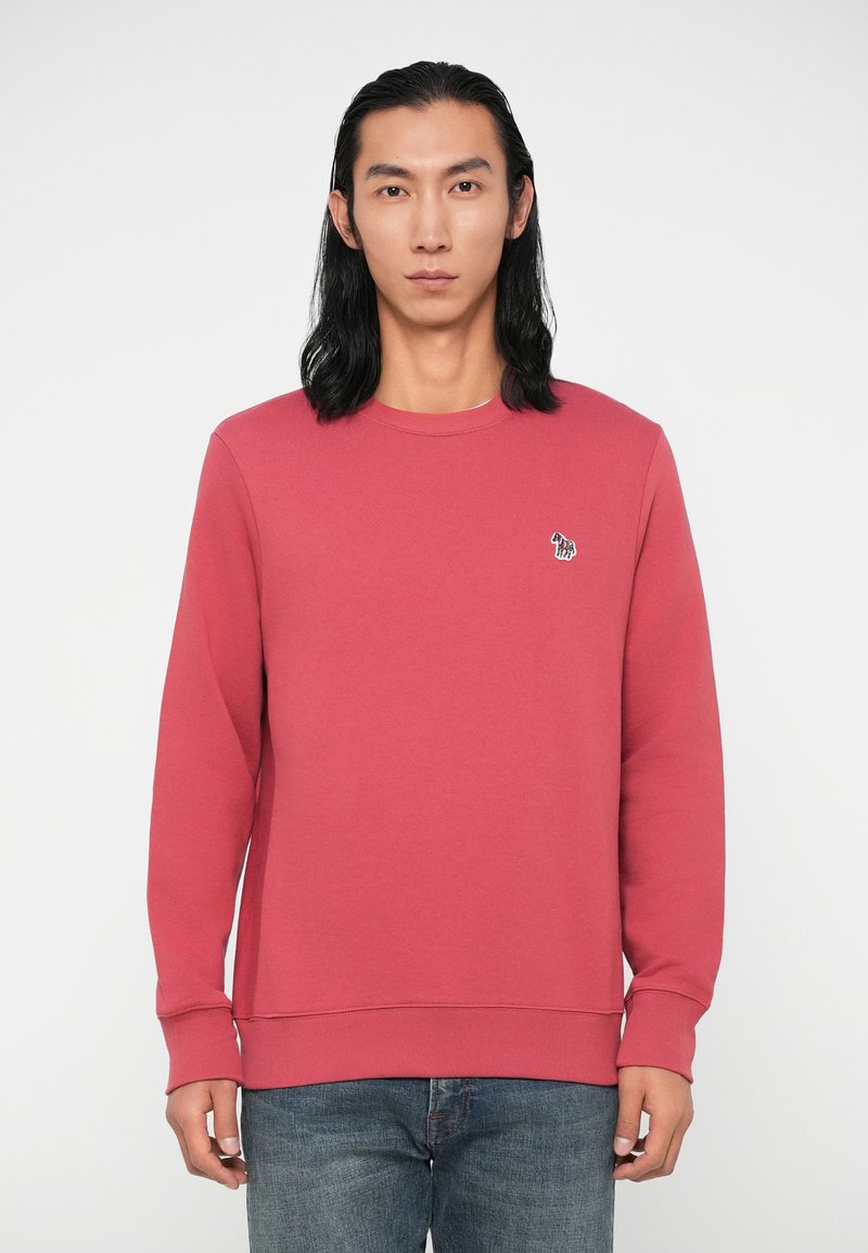 Raudonas džemperis, pagamintas iš minkštos medvilnės mišinio. Guma apykaklės ir apačios. Mažas siuvinėjimas kairėje krūtinės pusėje. Suderintas su mėlynomis džinsinėmis kelnėmis.