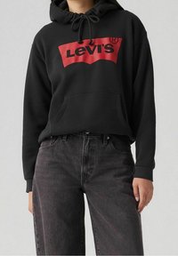 Sweat à capuche noir avec un logo Levi's rouge, doté d'une poche avant et d'une capuche avec cordon de serrage, associé à un jean gris foncé. Design simple et décontracté.
