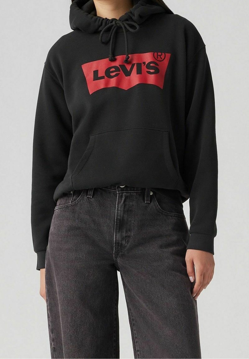 Sweat à capuche noir avec un logo Levi's rouge, doté d'une poche avant et d'une capuche avec cordon de serrage, associé à un jean gris foncé. Design simple et décontracté.