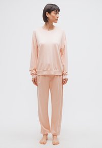 Γυναίκα που στέκεται ξυπόλυτη, φοράει αέρινο, μακρυμάνικο ροζ τοπ και παντελόνι στο ίδιο χρώμα, κοιτάζει προς τα δεξιά της πάνω σε λευκό, μονόχρωμο φόντο.