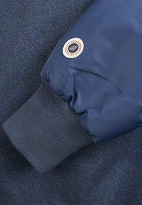 Veste bleu marine en matériau lisse et brillant avec poignets côtelés. Un patch circulaire avec un logo est cousu sur la manche.
