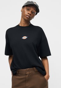 Νεαρός/ή που φοράει μαύρο μπλουζάκι Dickies, καφέ παντελόνι και σκούφο Dickies σκουρόγριου χρώματος, στέκεται με τα χέρια στις τσέπες μπροστά σε γκρι φόντο.