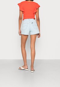 Shorts de mezclilla azul claro con dobladillo deshilachado, combinados con una blusa naranja de canalé con mangas acampanadas. La modelo lleva sandalias deslizantes beige.