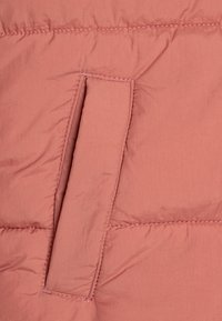 Vadderad jacka i mjuk rosaröd, med en rektangulär ficka och sömnadsdetaljer längs sömmarna. Slät och lätt tygtextur.