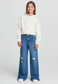 Felpa crema con zip frontale, polsini e orlo a coste, abbinata a jeans blu a gamba larga con dettagli strappati e orli sfilacciati.