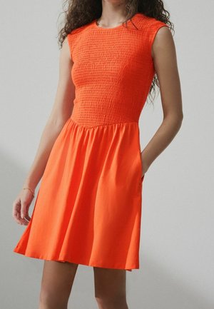 Femme portant une robe sans manches orange vif avec un corsage texturé et une jupe évasée, debout avec une main dans la poche sur un fond uni.