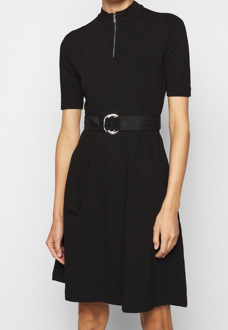 Schwarzes Kurzarmkleid mit hohem Kragen, Reißverschluss vorne, strukturiertem Stoff und einem breiten schwarzen Gürtel mit einer runden silbernen Schnalle.