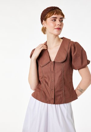 Woman wearing a brown buttoned blouse with puffed sleeves and wide collar, white skirt, and textured brown headband, looking right.