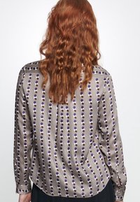 Långärmad, silkeslen blus i taupe med ett upprepat cirkulärt mönster i lila och brunt. Har knappade manschetter och en mjuk textur.