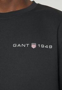 Chemise noire avec texte blanc « GANT 1949 » et un petit emblème en forme de bouclier avec des étoiles et des rayures sur la poitrine.