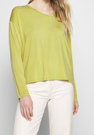 Femme portant un haut ample à manches longues vert citron associé à un pantalon blanc taille haute devant un fond uni.