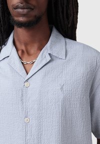 Camicia azzurra chiara con bottoni, realizzata in un tessuto testurizzato. Presenta un colletto a punta, due bottoni e un piccolo dettaglio ricamato sul petto.