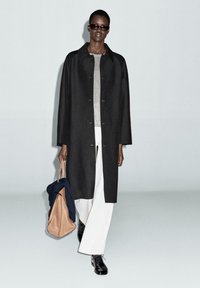 Cappotto lungo nero con colletto, maglia grigia e pantaloni bianchi. Borsa marrone con un oggetto blu, scarpe nere e occhiali da sole. Design minimalista.