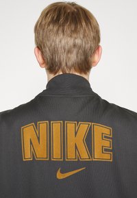 Svart jacka med en stor, iögonfallande, gyllen "NIKE"-logotyp och en swoosh. Tyget ser slätt ut med en strukturerad kragdesign.