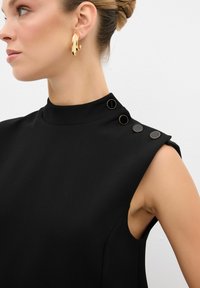 Prenda negra de cuello alto con hombros estructurados y tres botones decorativos. Tela suave, corte entallado y sin mangas. Pendientes de aro dorados.
