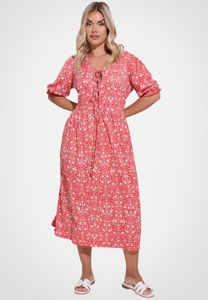 Kobieta stojąca z rękami na biodrach, ubrana w czerwoną sukienkę midi w kwiaty z bufkami na rękawach i białe sandały na płaskiej podeszwie.