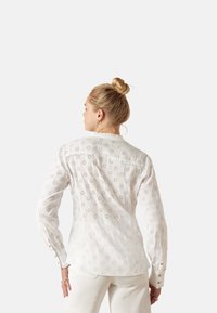 White long-sleeve shirt featuring floral eyelet pattern, buttoned cuffs, and a slightly curved hem. Soft, breathable fabric with a textured look.
