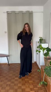 Sweatshirt preto com fecho éclair combinado com calças largas pretas. O modelo está em um chão de madeira, rodeado por plantas e paredes de cor clara.
