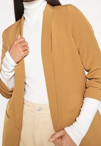 Femme portant un blazer camel sur un col roulé blanc et un pantalon beige, tenant un revers du blazer d'une main.