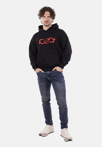 Felpa nera con un design di fiamme rosse, tasca frontale, abbinata a jeans blu e sneaker bianche. Sfondo bianco uniforme.