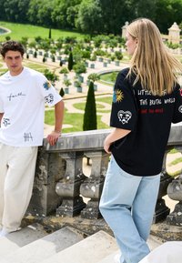 Deux personnes portant des t-shirts graphiques se tiennent sur des marches en pierre surplombant un jardin formel avec des plantes vertes et des allées.