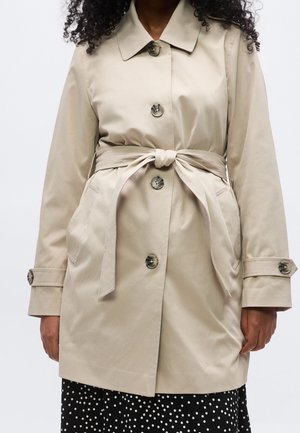 Femme portant un trench-coat beige ceinturé avec de gros boutons sur une jupe noire à pois blancs sur fond uni.