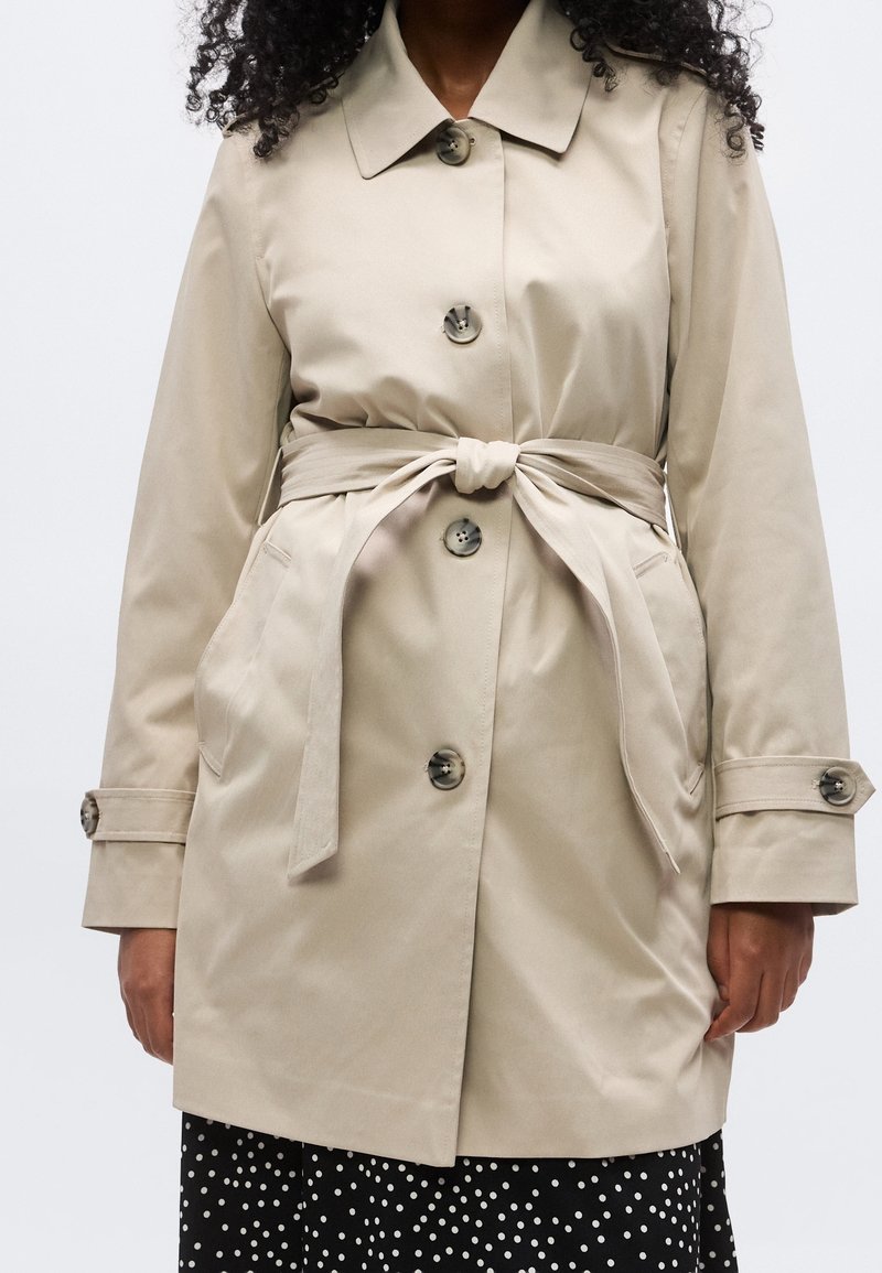 Femme portant un trench-coat beige ceinturé avec de gros boutons sur une jupe noire à pois blancs sur fond uni.