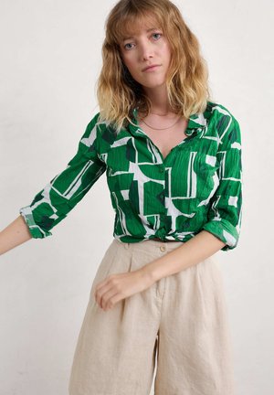 Camisa de botones verde y blanca con estampado, con mangas enrolladas, combinada con pantalones de tiro alto beige que tienen un botón frontal y pliegues.