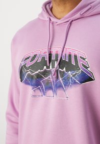 Ανοιχτό ροζ φούτερ με κουκούλα που διαθέτει σχεδιασμό γραφικών με τη λέξη "FORTNITE" σε μαύρο και μωβ χρώμα, συνοδευόμενο από διακριτά σχέδια αστραπών και βουνών.