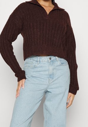 Femme portant un pull court côtelé marron foncé avec un col et un jean taille haute bleu clair, main reposant sur la hanche.