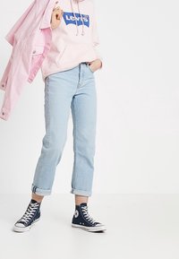Sweat à capuche Levi's rose clair avec logo bleu, associé à un jean bleu clair retroussé et des baskets Converse navy. Veste rose tenue à la main gauche.