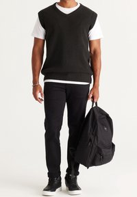 Gilet nero lavorato a maglia con scollatura a V sopra una maglietta bianca, abbinato a pantaloni neri e uno zaino nero. Sneakers nere completano l'outfit.