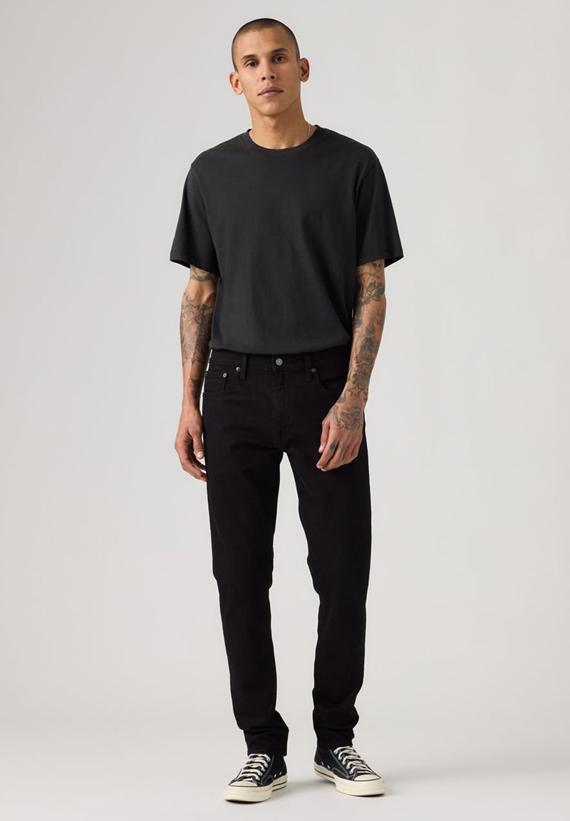 Jeune homme tatoué portant un t-shirt noir, un jean noir et des baskets noires, debout devant un fond clair uni.