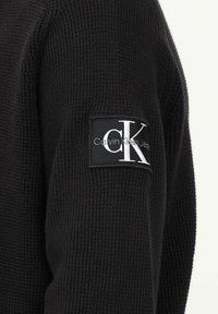 Maglione nero in maglia con tessuto testurizzato, dotato di una toppa rettangolare nera sulla manica che presenta il logo "Calvin Klein Jeans" in bianco.