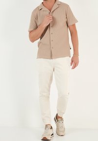 Ljusbrun kortärmad skjorta med knappar i en texturerad tyg, parat med vita joggerbyxor och beige sneakers. Enkelt, avslappnat designs.