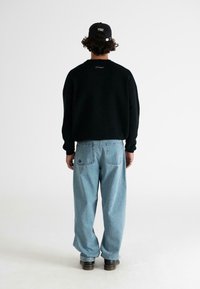 Persona con capelli ricci che indossa un cappello nero, un maglione nero, jeans larghi di un blu chiaro e stivali neri, in piedi di spalle contro uno sfondo bianco.